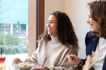 Deux collègues souriantes partagent un repas dans un restaurant, assises à une table près d’une fenêtre.