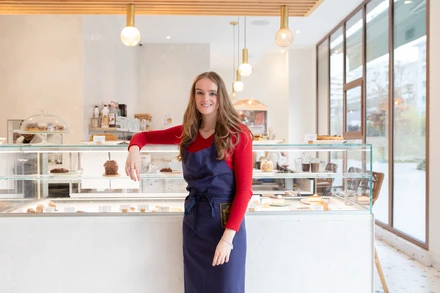 Une employée souriante en tablier se tient derrière la vitrine d’une boulangerie présentant des pâtisseries.
